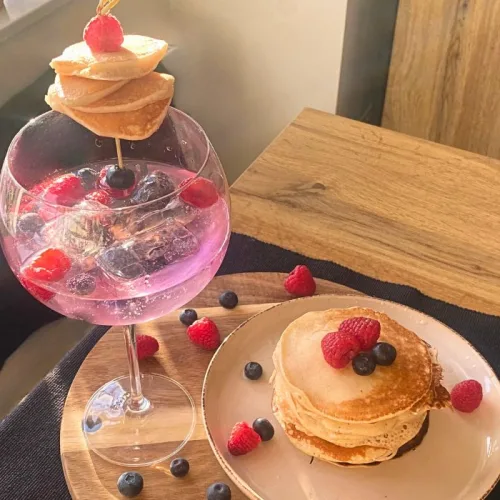 gin and tonic in a copa glass filled with large ice cubes and berries, garnished with a mini pancake stack. Large stack of pancakes covered in a G&T syrup with fresh berries.