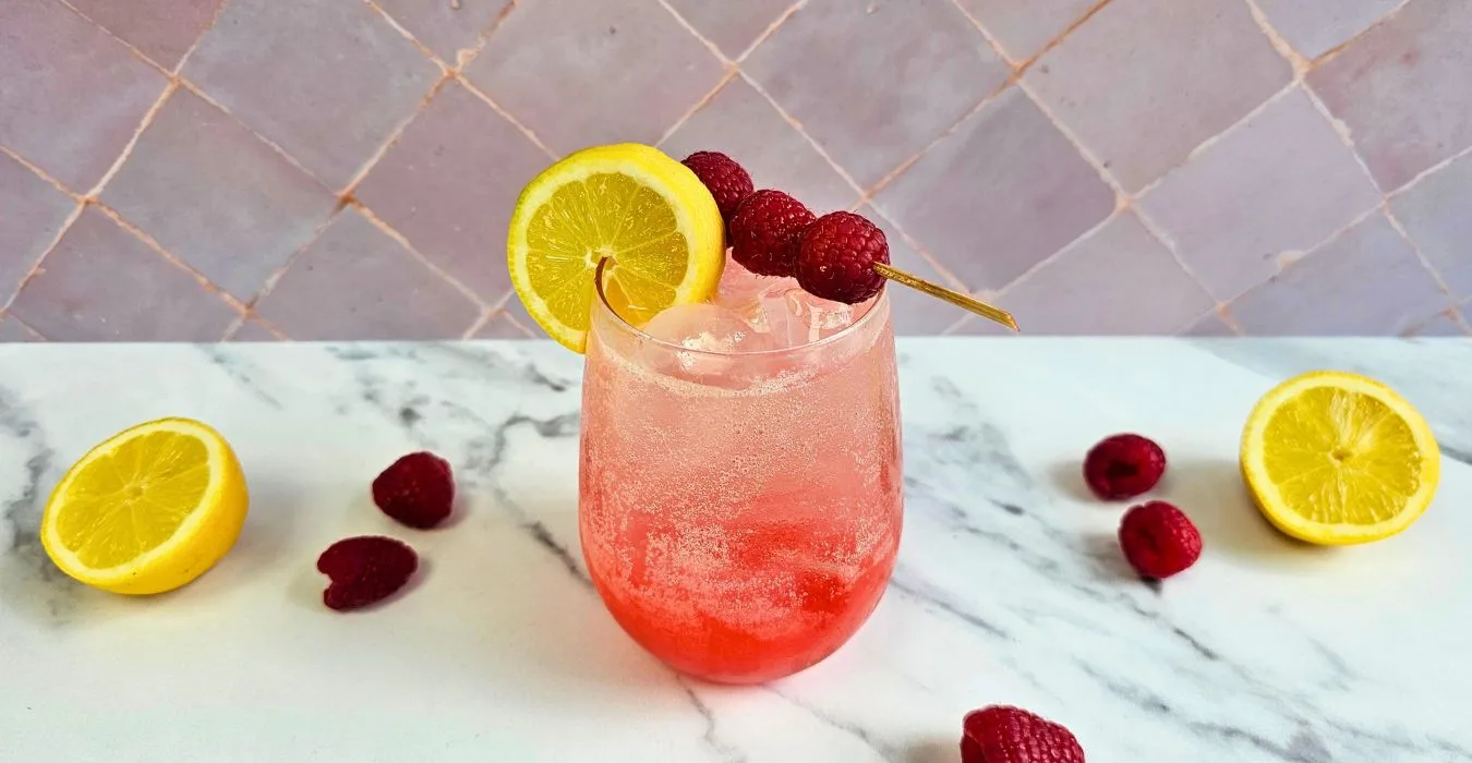 Limoncello berry burst cocktail on a white marble counter surroundd by fresh raspberries and lemon.