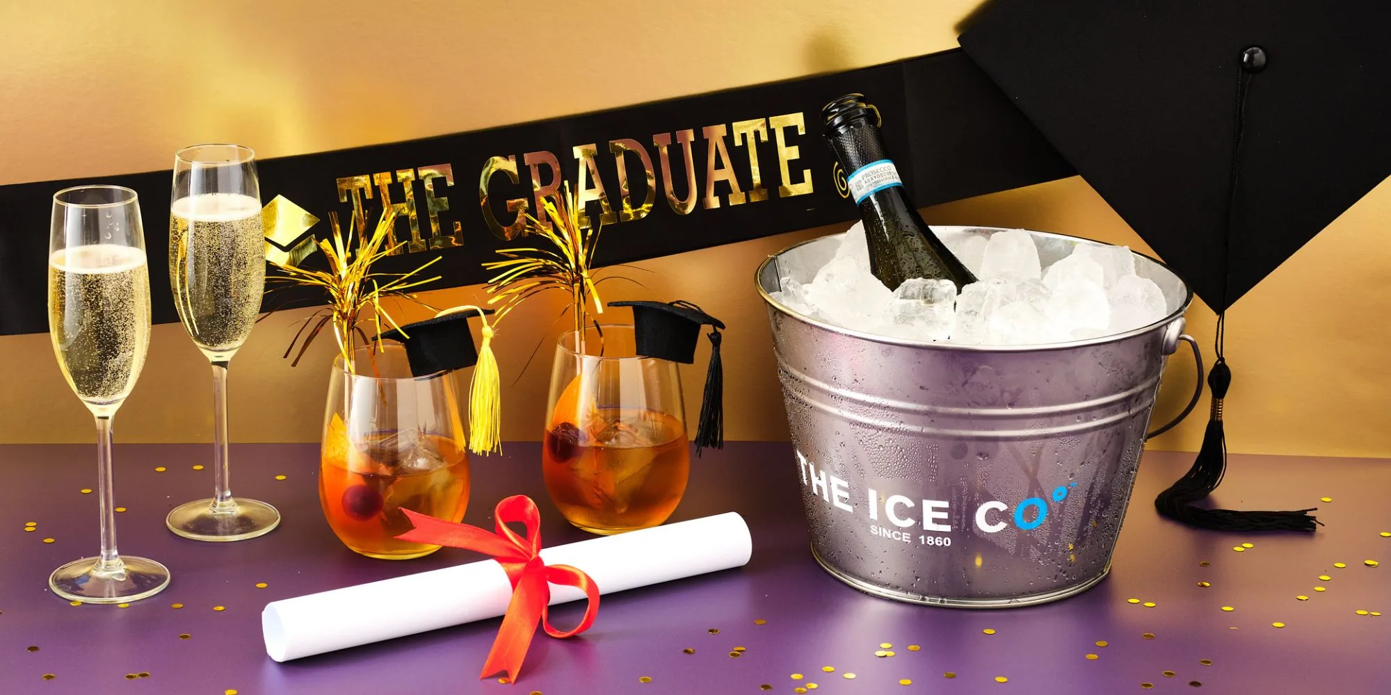graduation cap and sash next to a bottle of prosecco chilling in and ice bucket and old fashioned cocktails