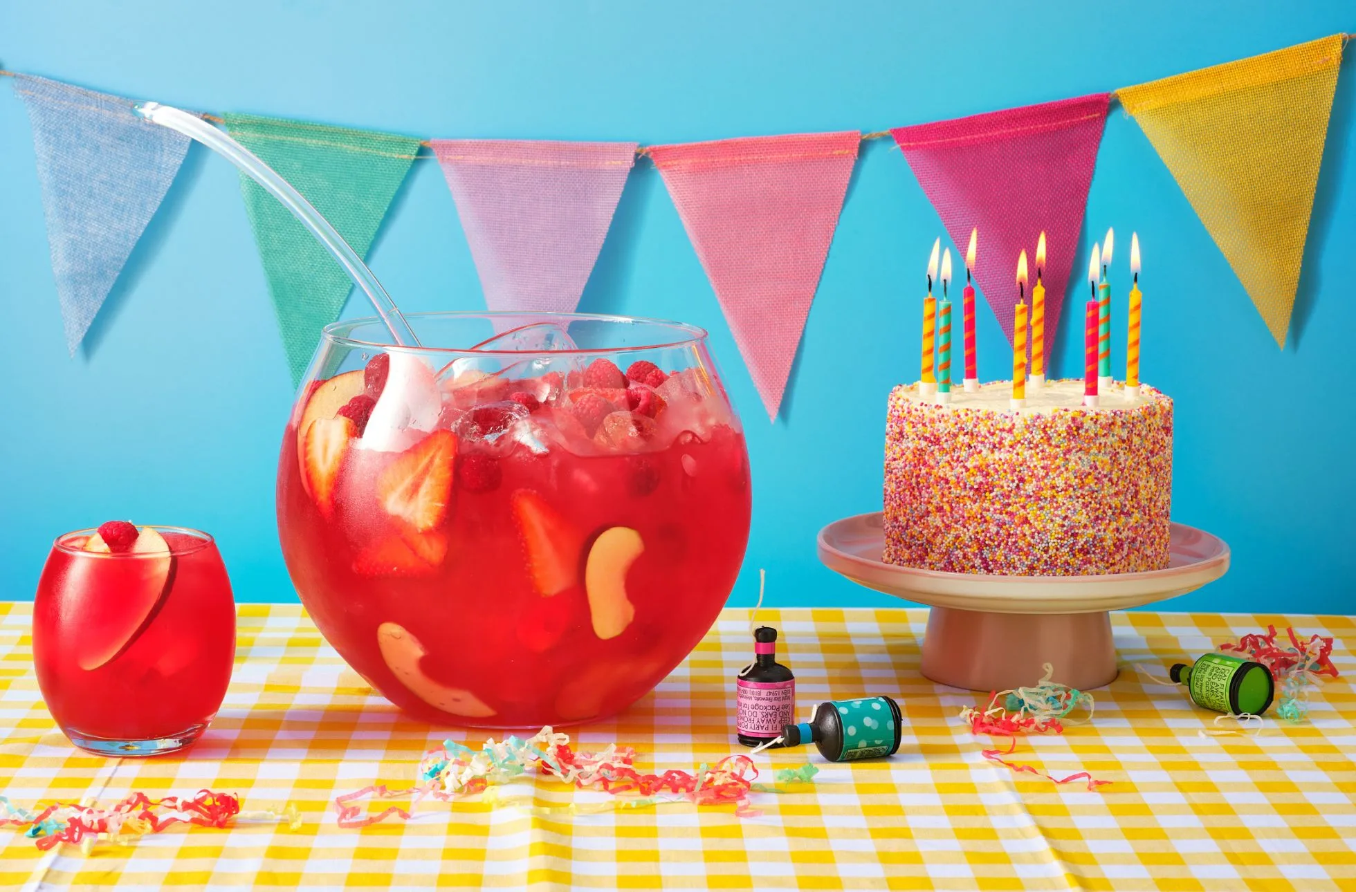 pastel bunting over a fruity fishbowl cocktail next to a sprinkle birthday cake with candles