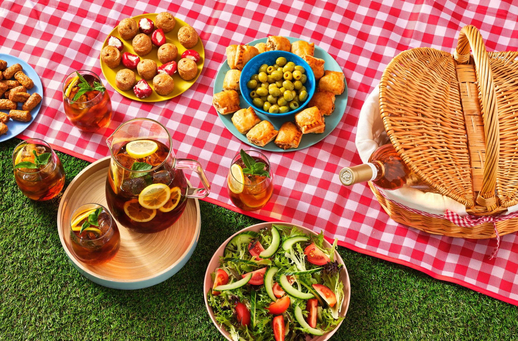 picnic blanket and hamper with rose wine, surrounded by picnic snacks and olives with a jug of refreshing iced tea