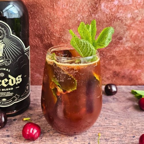 original deeds cherry smash cocktail next to a bottle of rum surrounded by cherries and mint