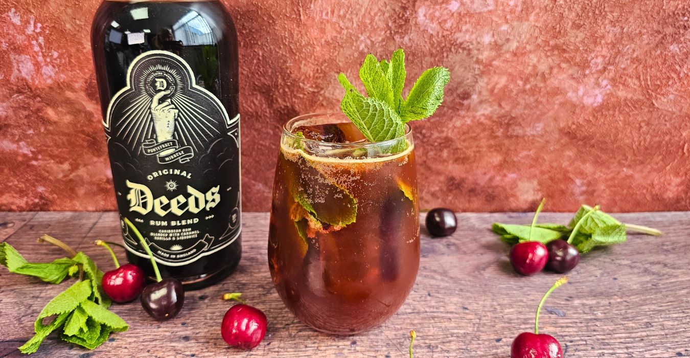 original deeds cherry smash cocktail next to a bottle of rum surrounded by cherries and mint