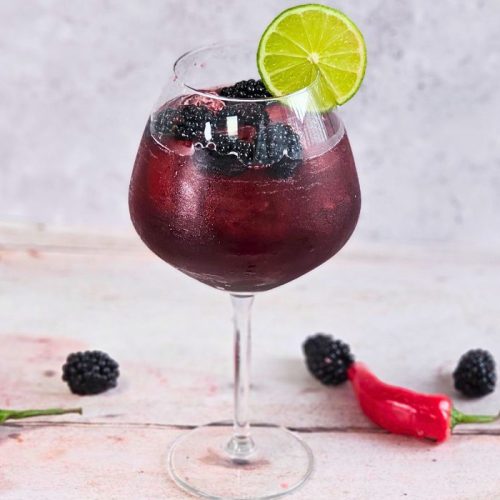 Hot Honey Blackberry Margarita against a plain background surrounded by berries and chillies, in a large gin glass packed with Party Ice