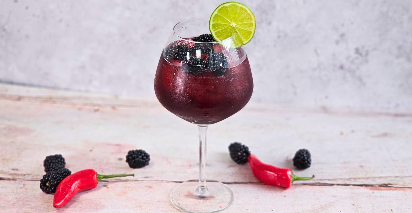 Hot Honey Blackberry Margarita against a plain background surrounded by berries and chillies, in a large gin glass packed with Party Ice