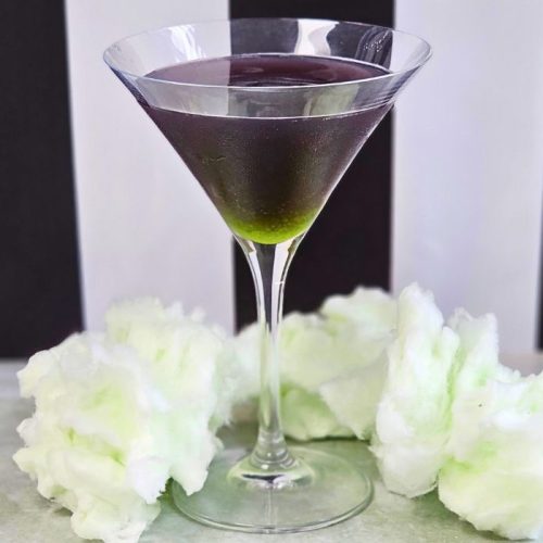 Purple Cocktail with green in the bottom against the iconic beetlejuice black and white stripy background