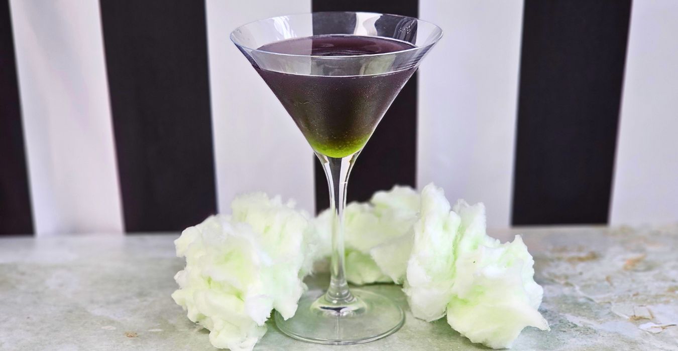 Purple Cocktail with green in the bottom against the iconic beetlejuice black and white stripy background